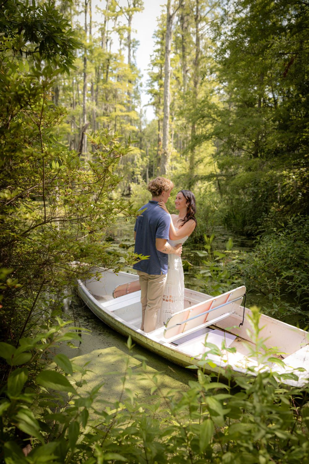 Couple standing and embracing in a small boat surrounded by lush green forest and calm water.