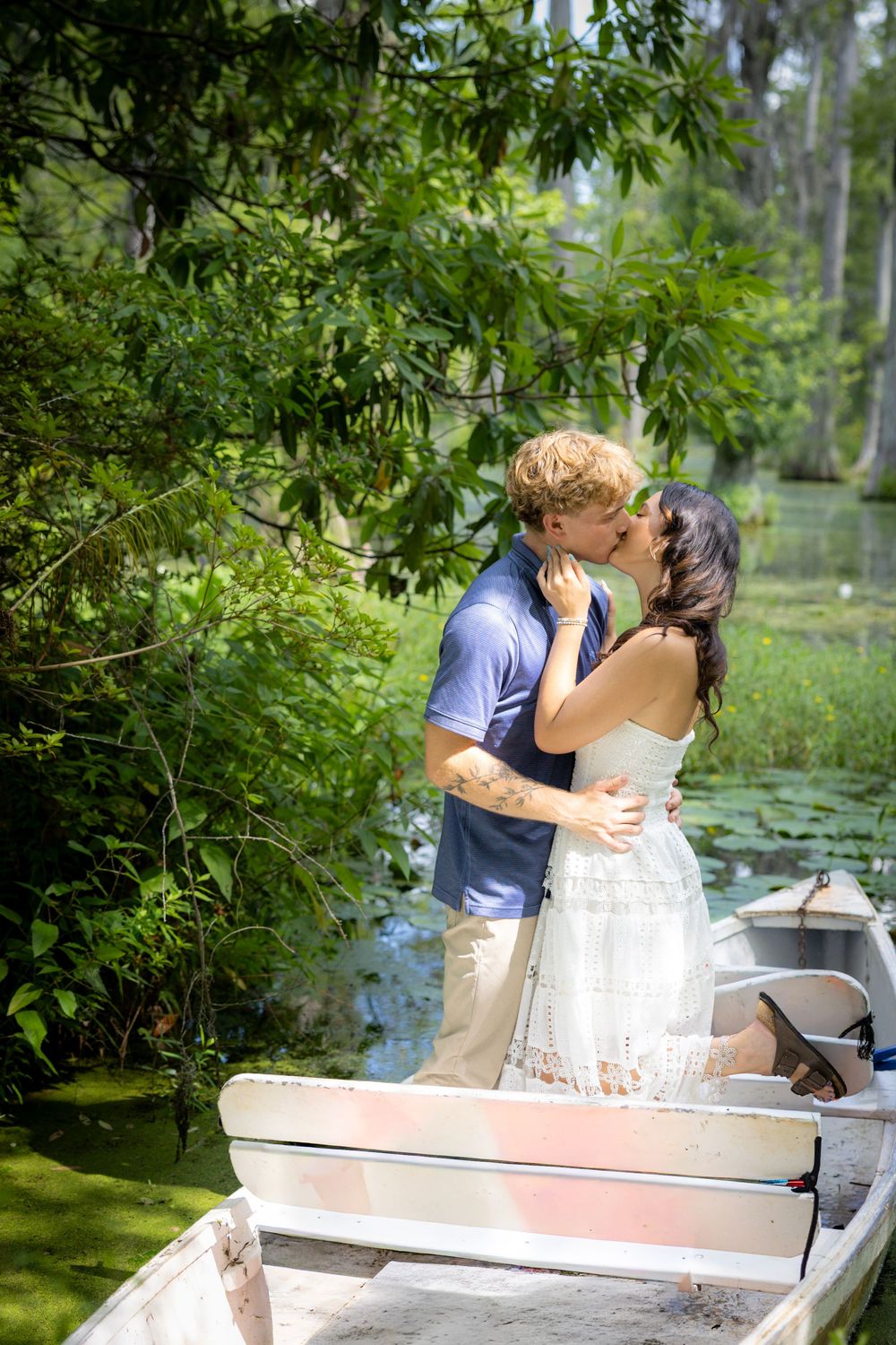 Couple sharing a romantic kiss in a white rowboat surrounded by lush green trees near a serene pond.