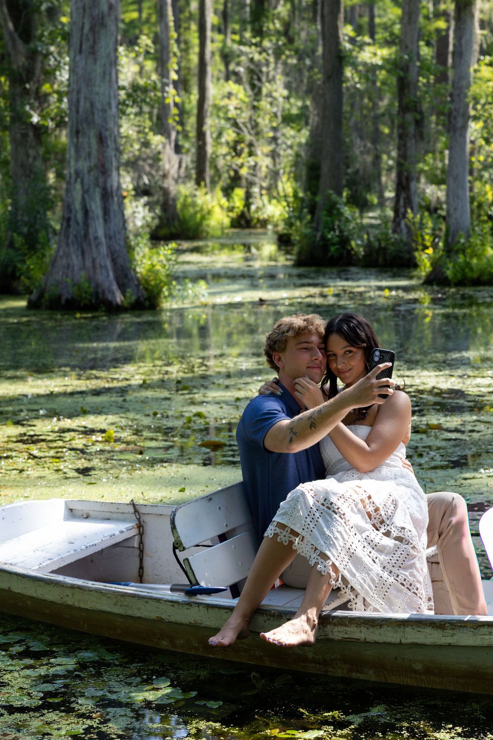 Couple taking a selfie in a rowboat surrounded by cypress trees and lily pads in a lush green swamp.