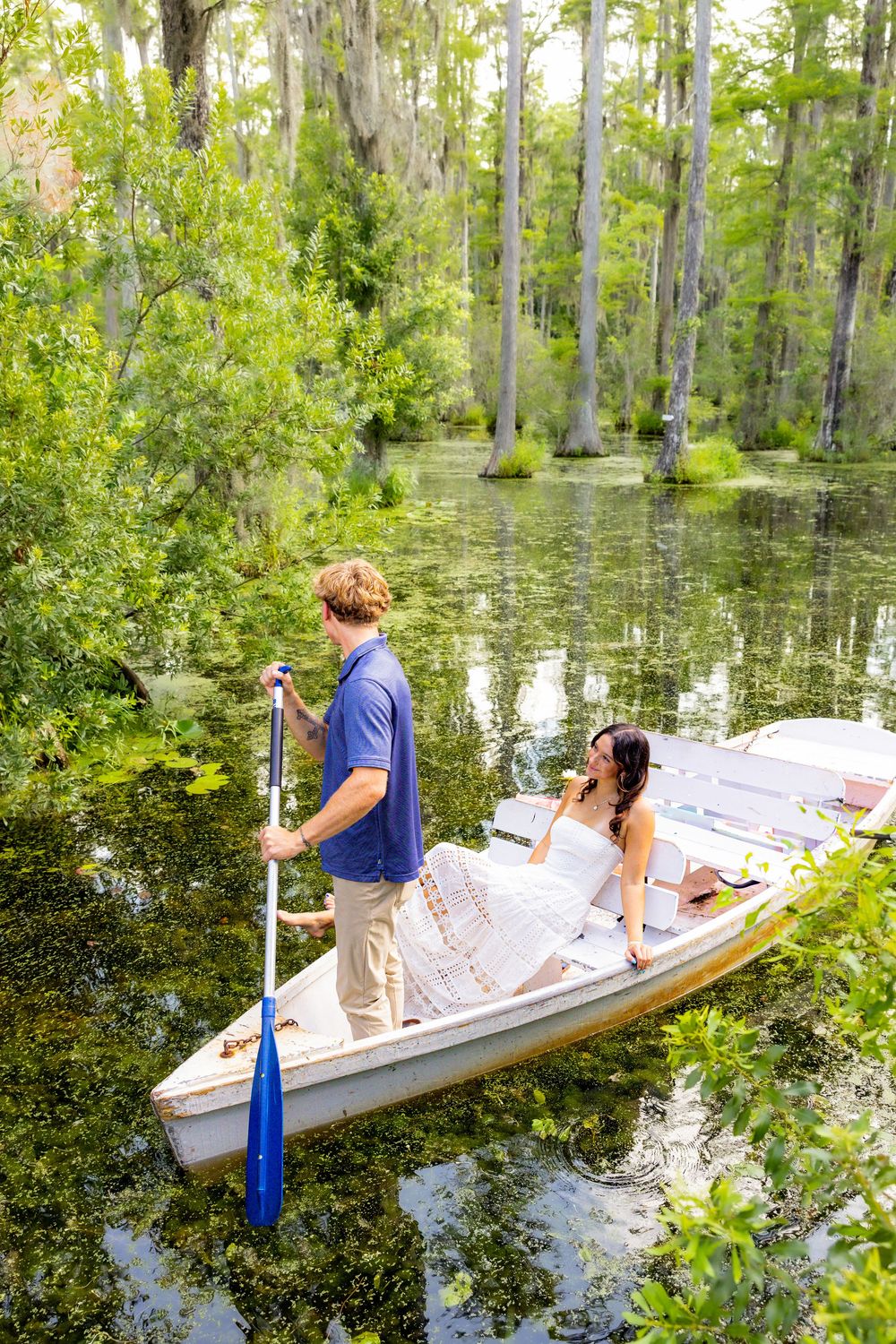 Couple on a rowboat in a lush green swamp, man standing with oar, woman seated in white dress.