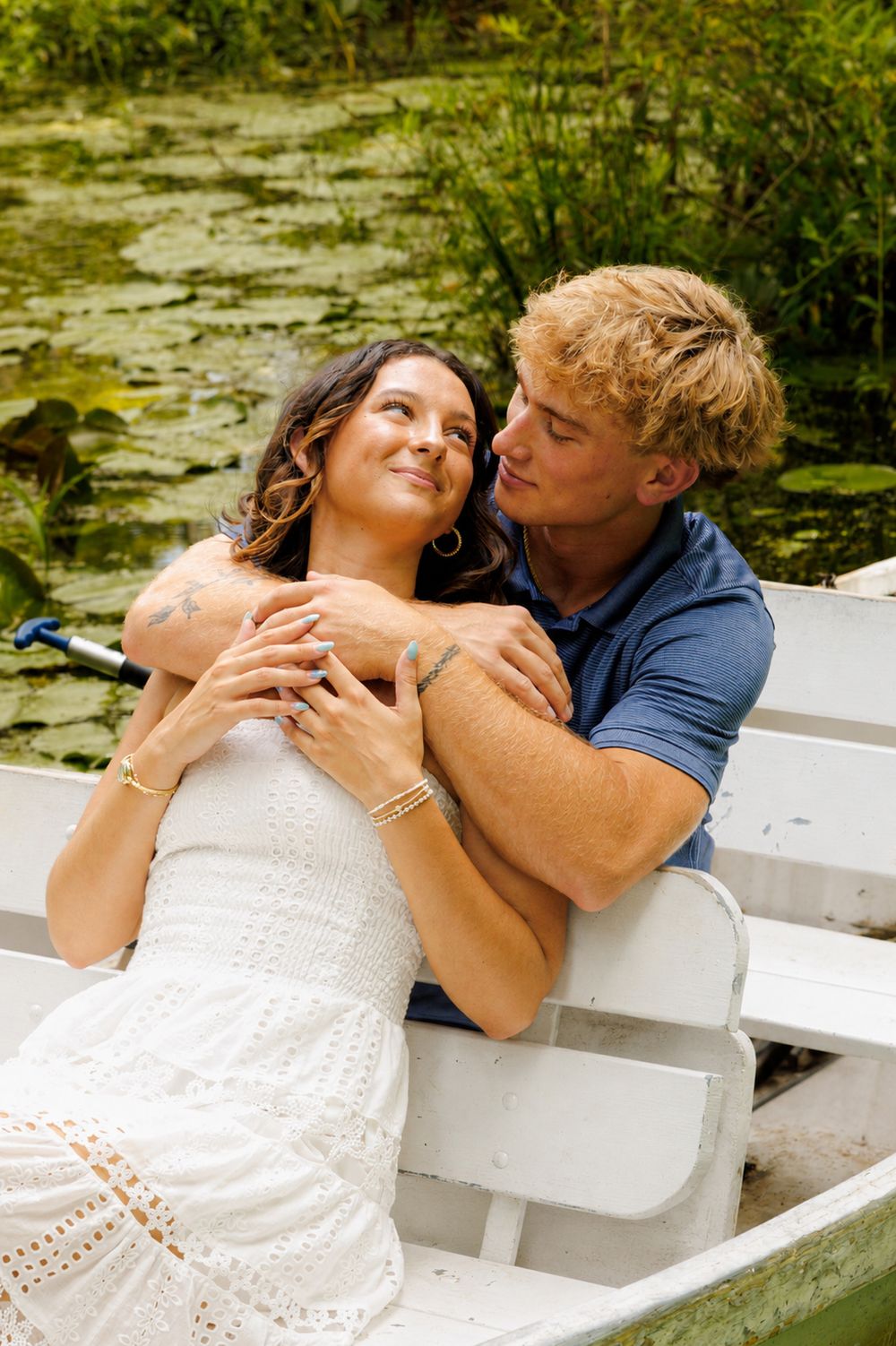 Couple embracing romantically in a white rowboat on a lily pad pond, woman in white dress gazing up at blond man.