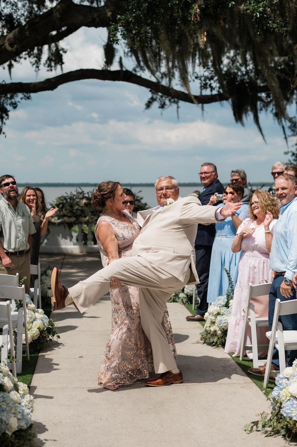 Bride and groom share a romantic dip kiss at outdoor waterfront wedding ceremony with guests applauding.