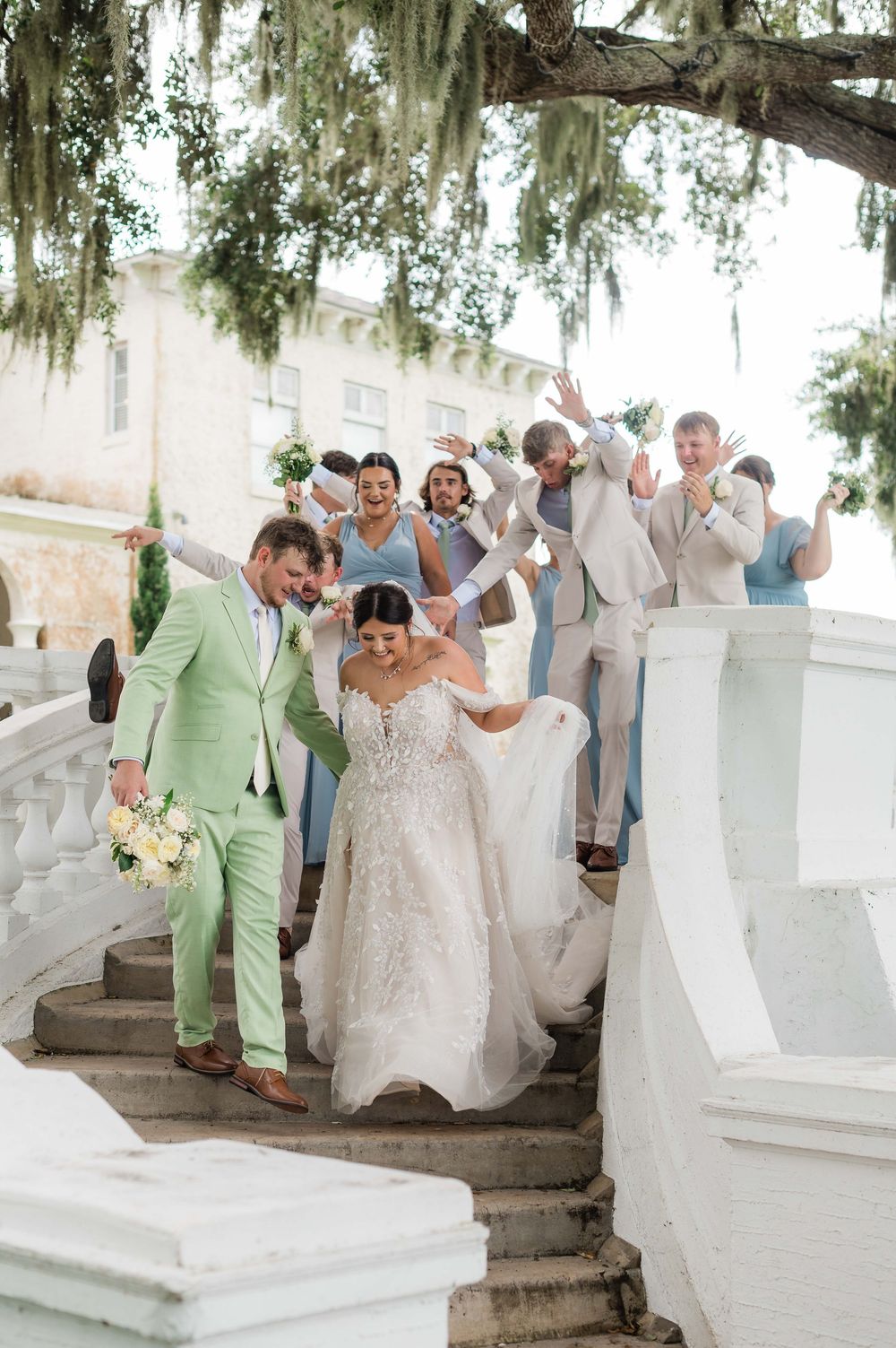 Jubilant wedding party celebrates on grand white staircase under Spanish moss-draped oak tree.