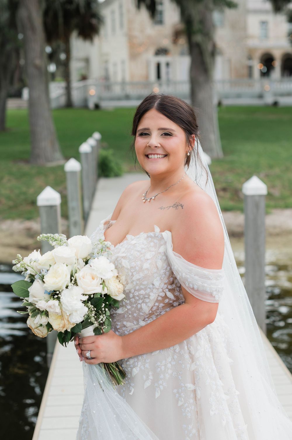 Smiling bride in off-shoulder lace gown holds white bouquet on dock near elegant venue.