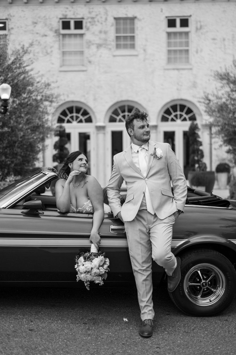 Black and white photo of bride and groom posing with vintage convertible outside elegant estate.