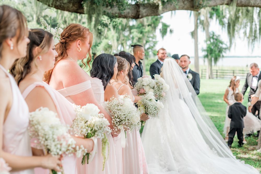 Bridesmaids in blush pink dresses holding baby's breath bouquets stand beside a bride in a white gown at an outdoor wedding.