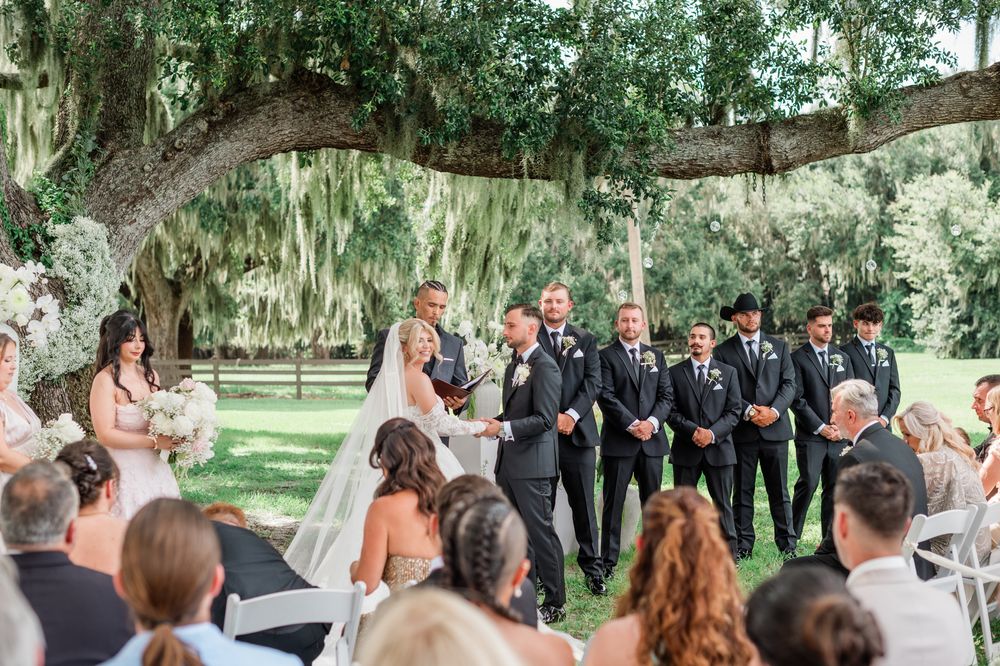 Wedding ceremony under large oak tree with bride, groom, and groomsmen standing at outdoor altar.