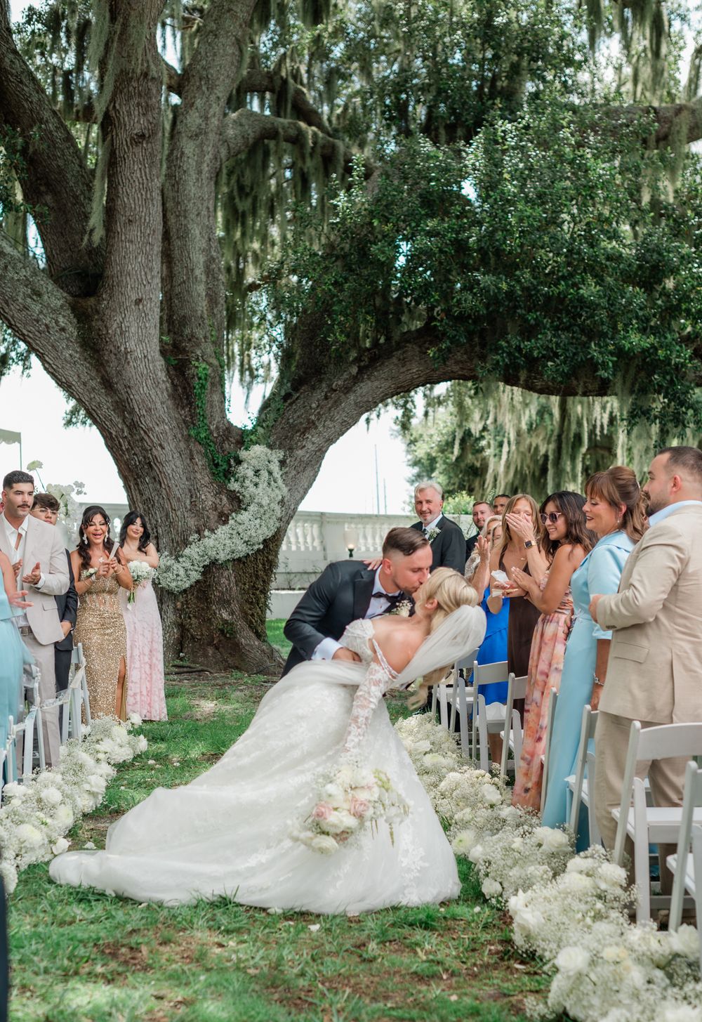 Groom dips bride in stunning lace gown at outdoor ceremony aisle lined with white floral arrangements.