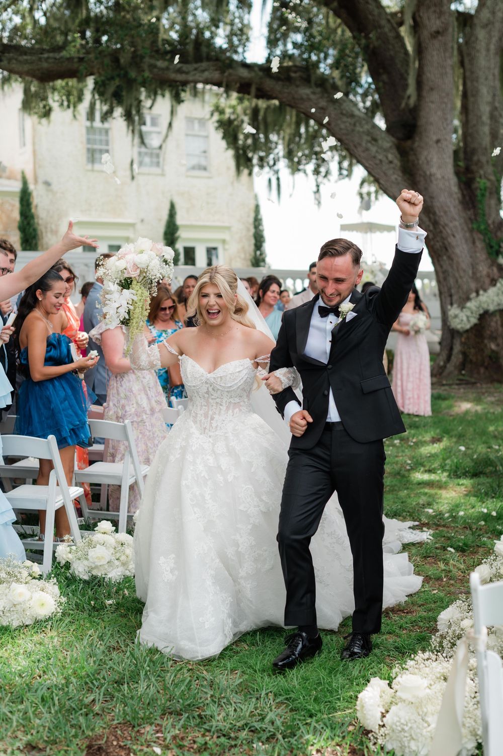 Joyful bride and groom celebrate walking back down the aisle after outdoor wedding ceremony under large oak tree.
