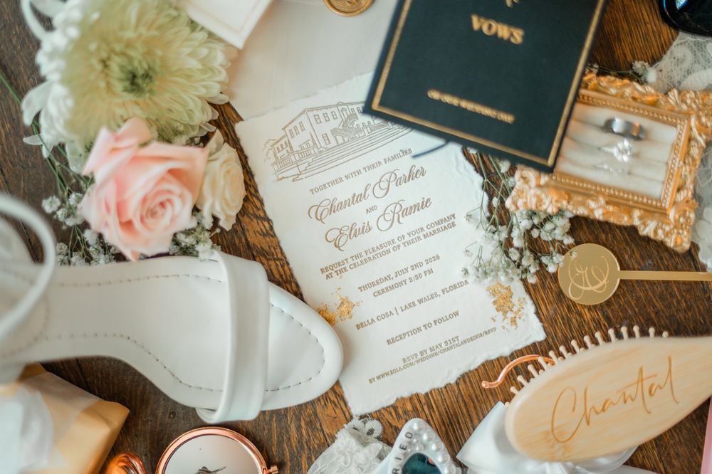 Elegant wedding flat lay with floral invitation, vow booklet, bridal heels, pink roses, and gold accessories on wood.