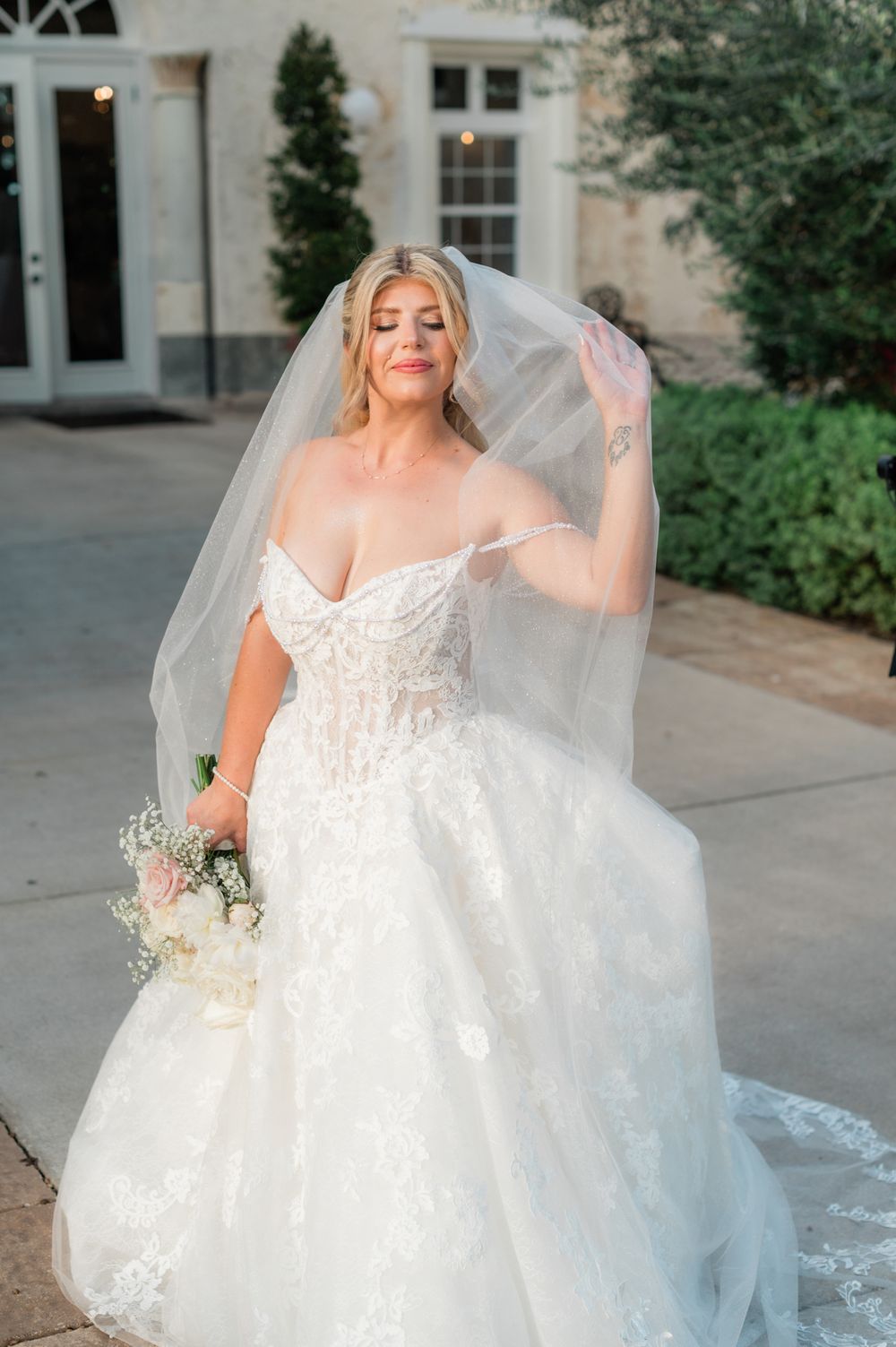Bride in off-shoulder lace ballgown lifts veil while holding white bouquet outside elegant venue.
