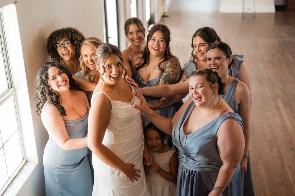 Bride in white gown poses with bridesmaids in dusty blue dresses, smiling together near a bright window.