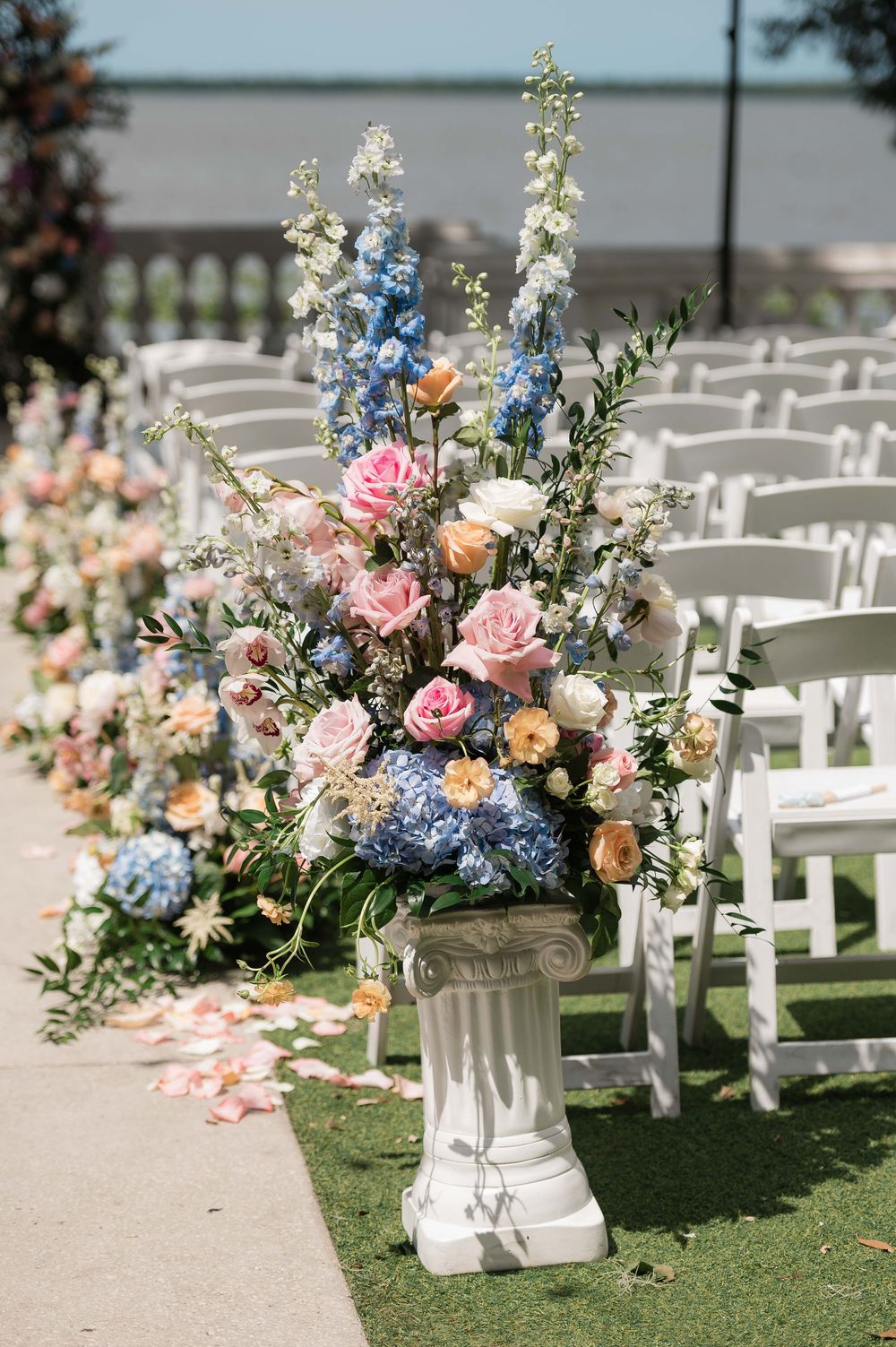 Elegant wedding ceremony aisle with white pedestal floral arrangement featuring pink roses, blue hydrangeas, and delphinium.