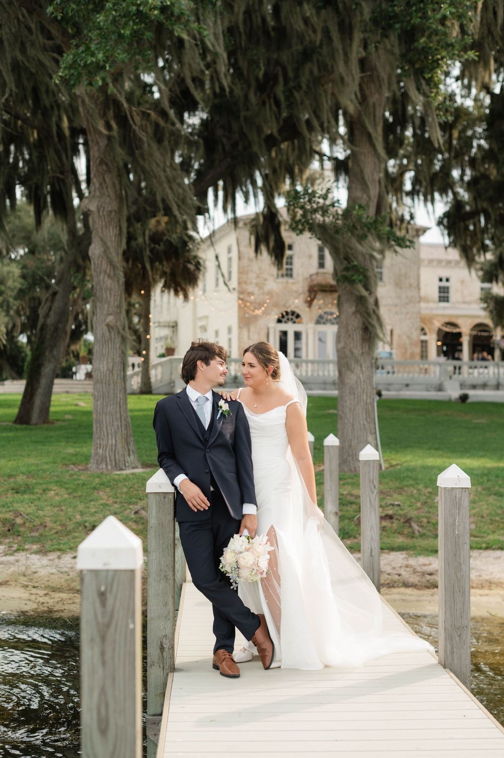 Bride and groom share a romantic moment on a dock, surrounded by moss-draped trees and an elegant estate.