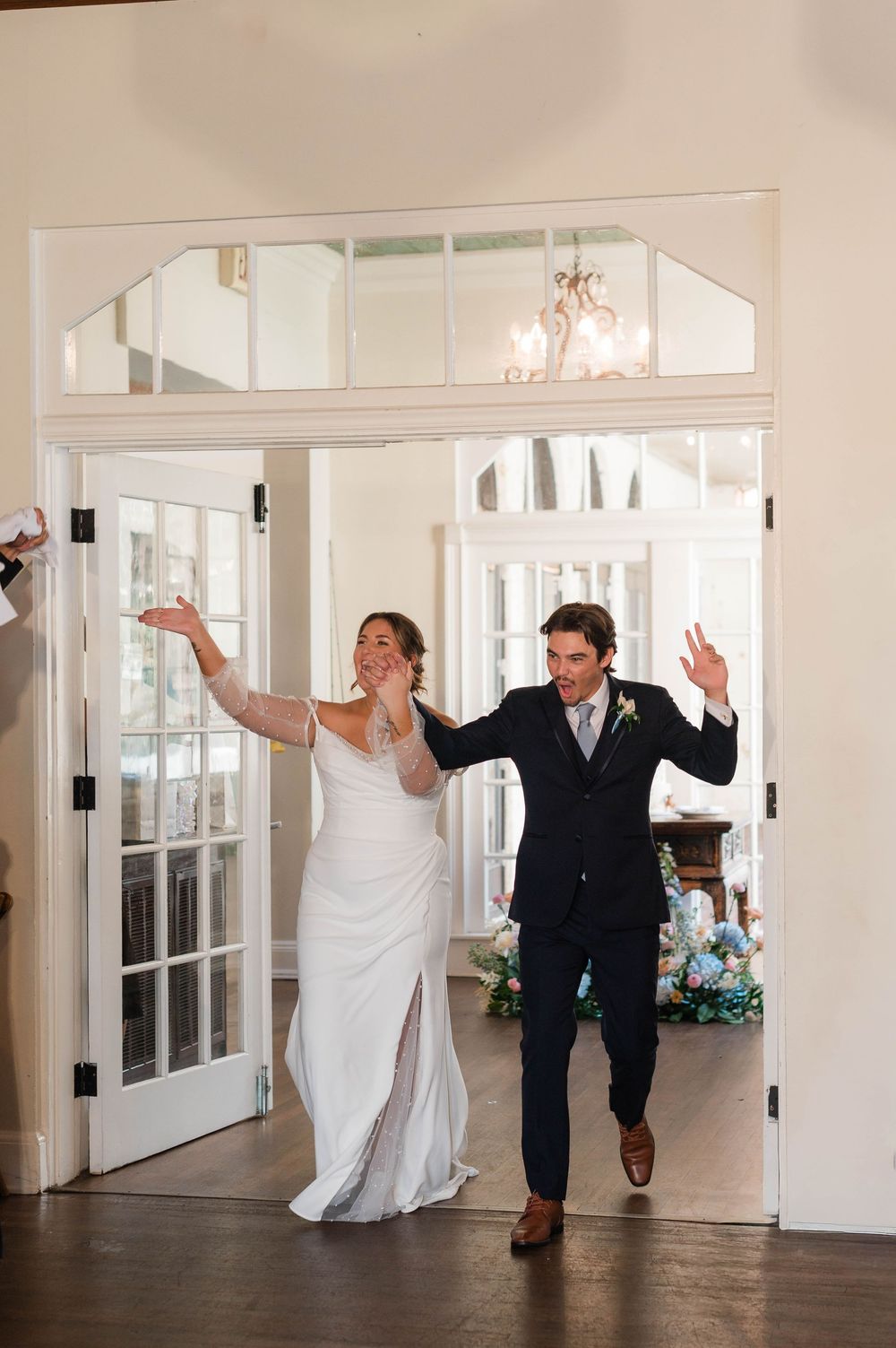 Bride and groom make grand reception entrance through French doors with arms raised in celebration.