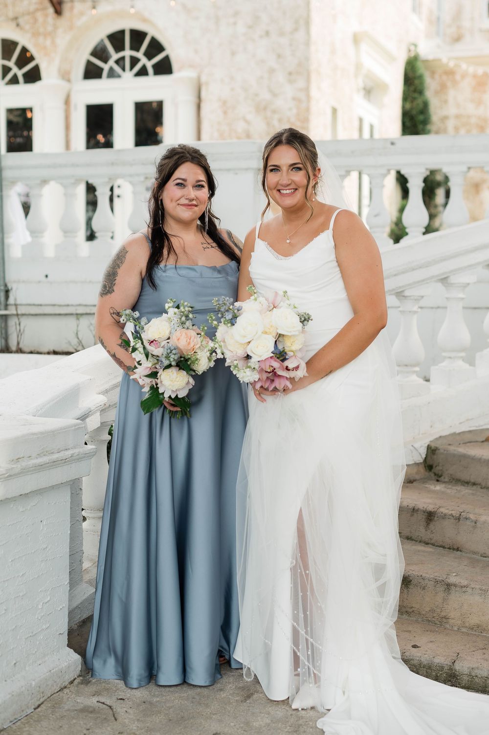 Bride in white gown poses with bridesmaid in dusty blue dress, both holding floral bouquets on elegant venue steps.