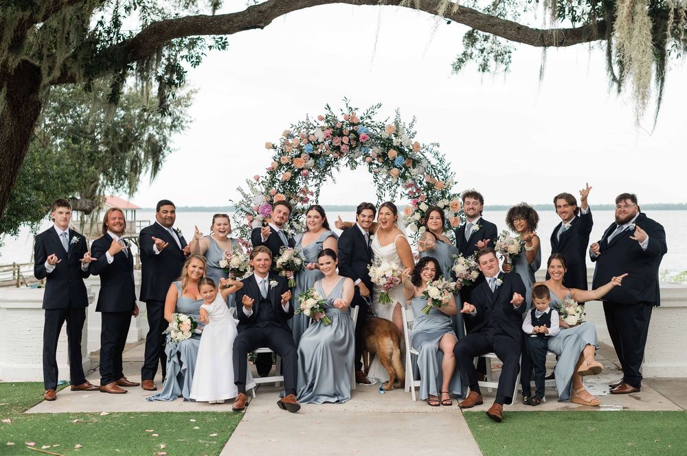 Large wedding party celebrates with champagne under Spanish moss by floral arch near waterfront.