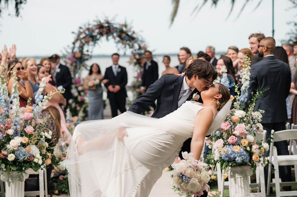 Groom dips bride for romantic kiss at outdoor wedding ceremony with floral arch backdrop.