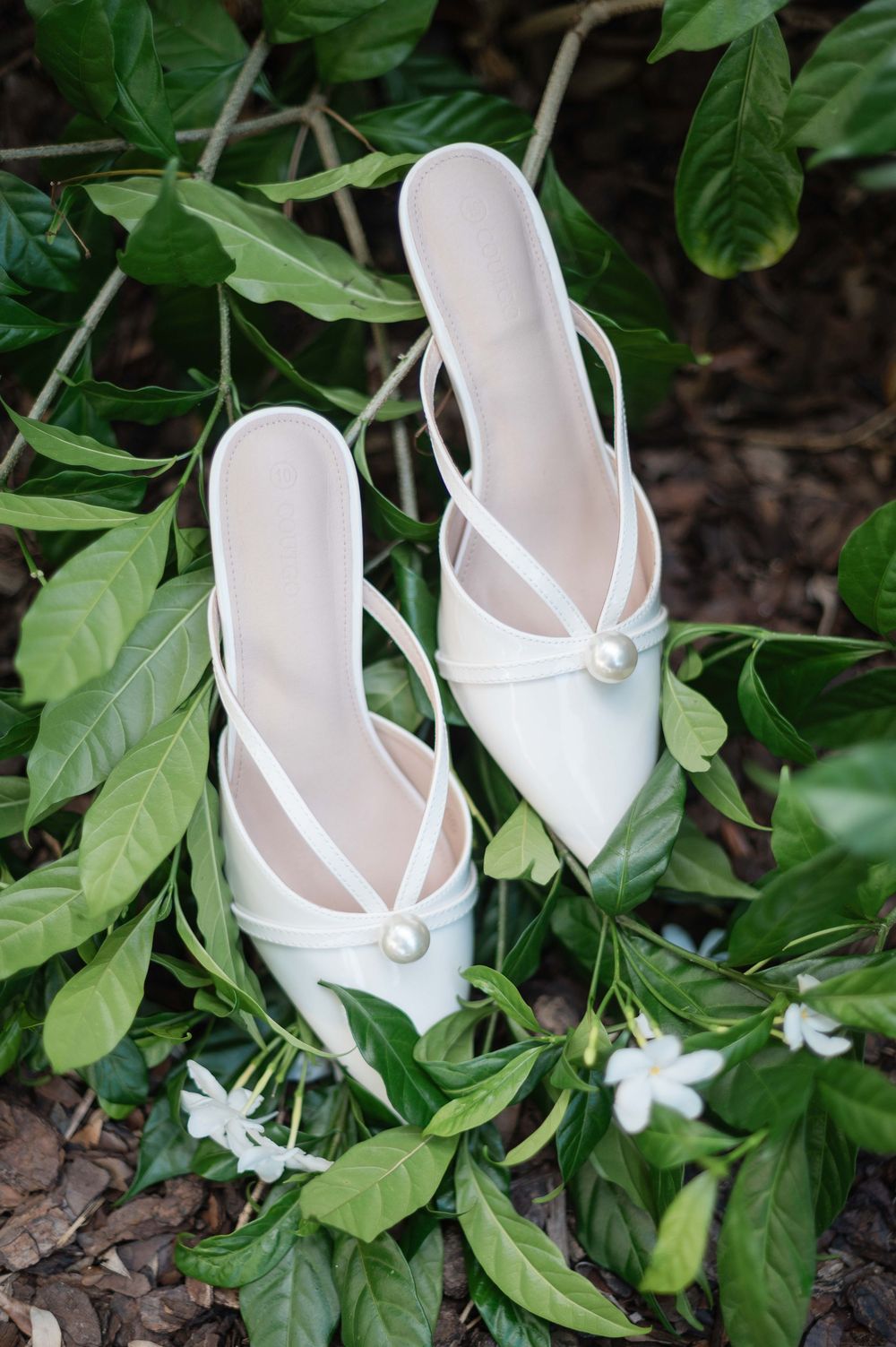 Elegant white pointed-toe mule heels with pearl detail resting on green leaves outdoors.