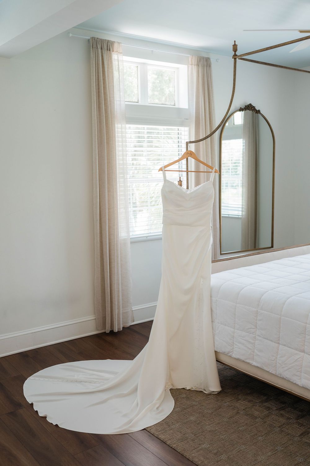 Elegant white wedding gown with train displayed on wooden hanger in bright bedroom with mirror and canopy bed.