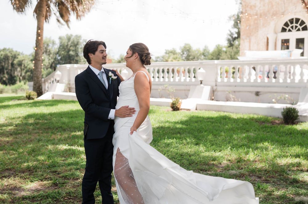 Bride and groom share tender moment on lush green lawn beside elegant white estate.