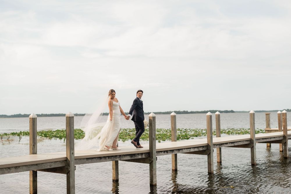 Bride and groom holding hands walking on a wooden dock over calm water on their wedding day.