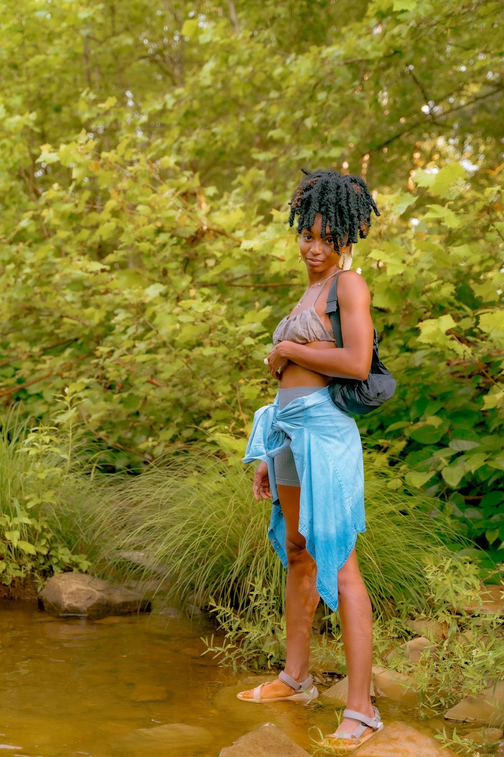 A person standing in a creek wearing casual summer attire against a backdrop of vibrant green foliage.