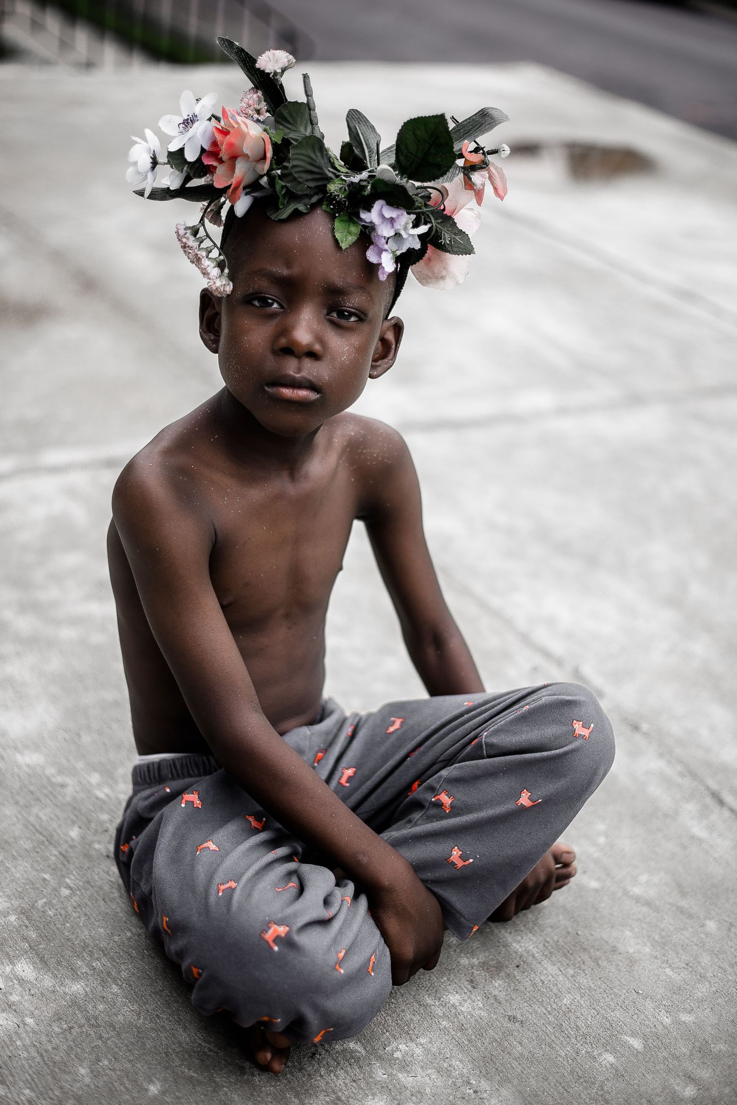 Child wearing floral headpiece and patterned pants sits cross-legged on concrete ground.