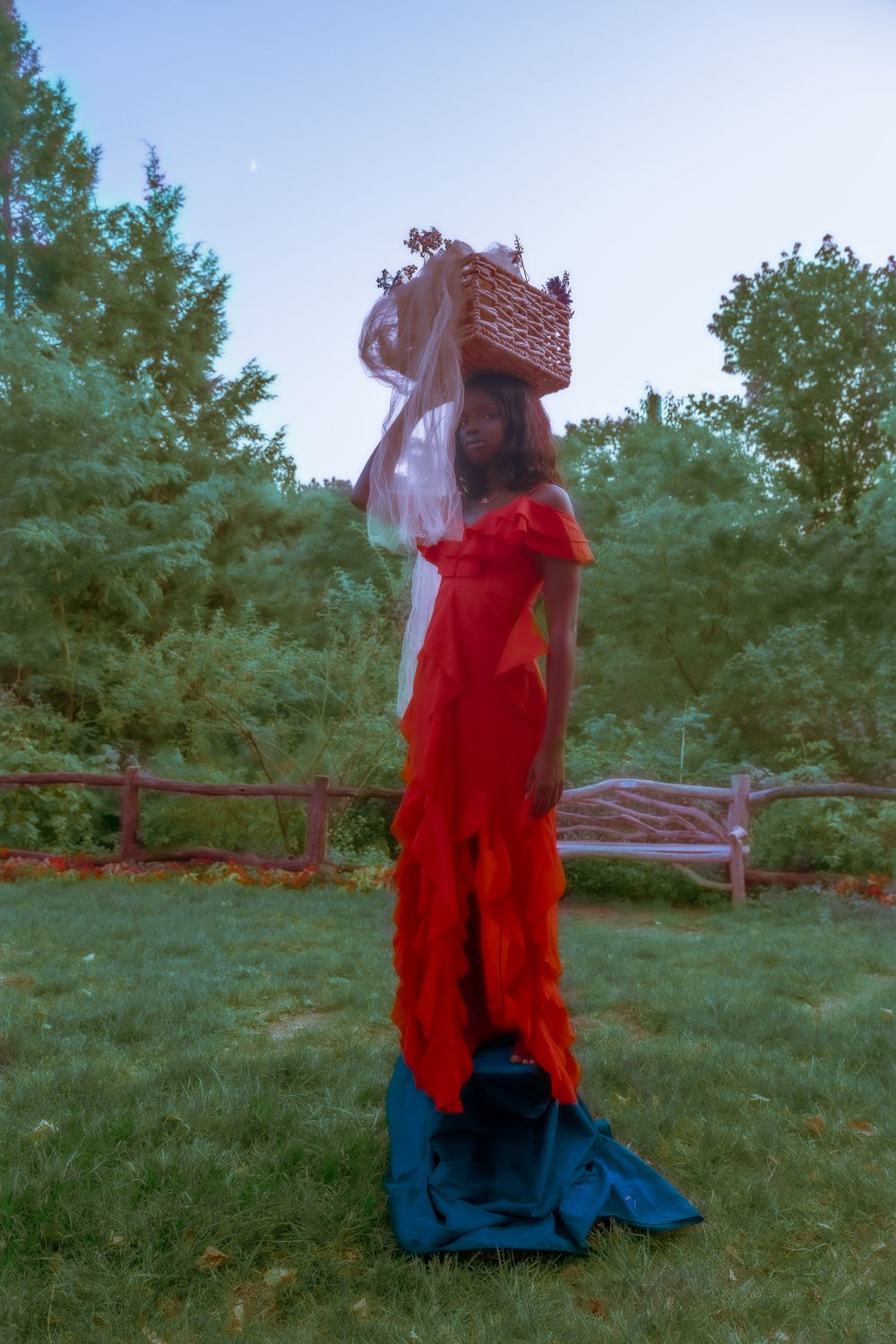 Figure in flowing red dress and headpiece stands in garden setting at dusk.