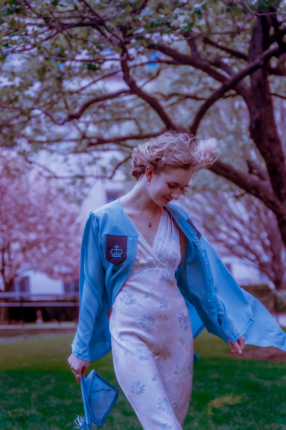 A dreamy portrait of someone in a blue jacket and floral dress standing beneath cherry blossom trees.