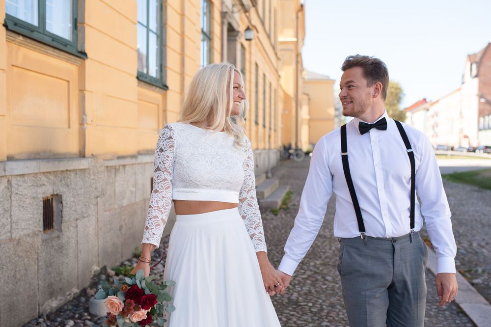 A couple in wedding attire walking hand in hand along a European street with historic architecture.