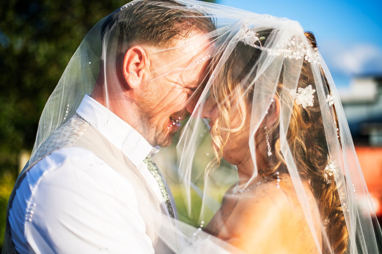 Bride and groom share an intimate moment under a flowing white wedding veil, laughing together in golden sunlight.