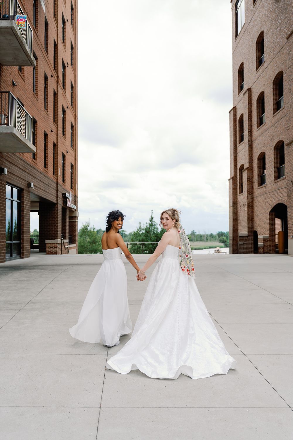 Two brides in white gowns hold hands walking between brick buildings on their wedding day.