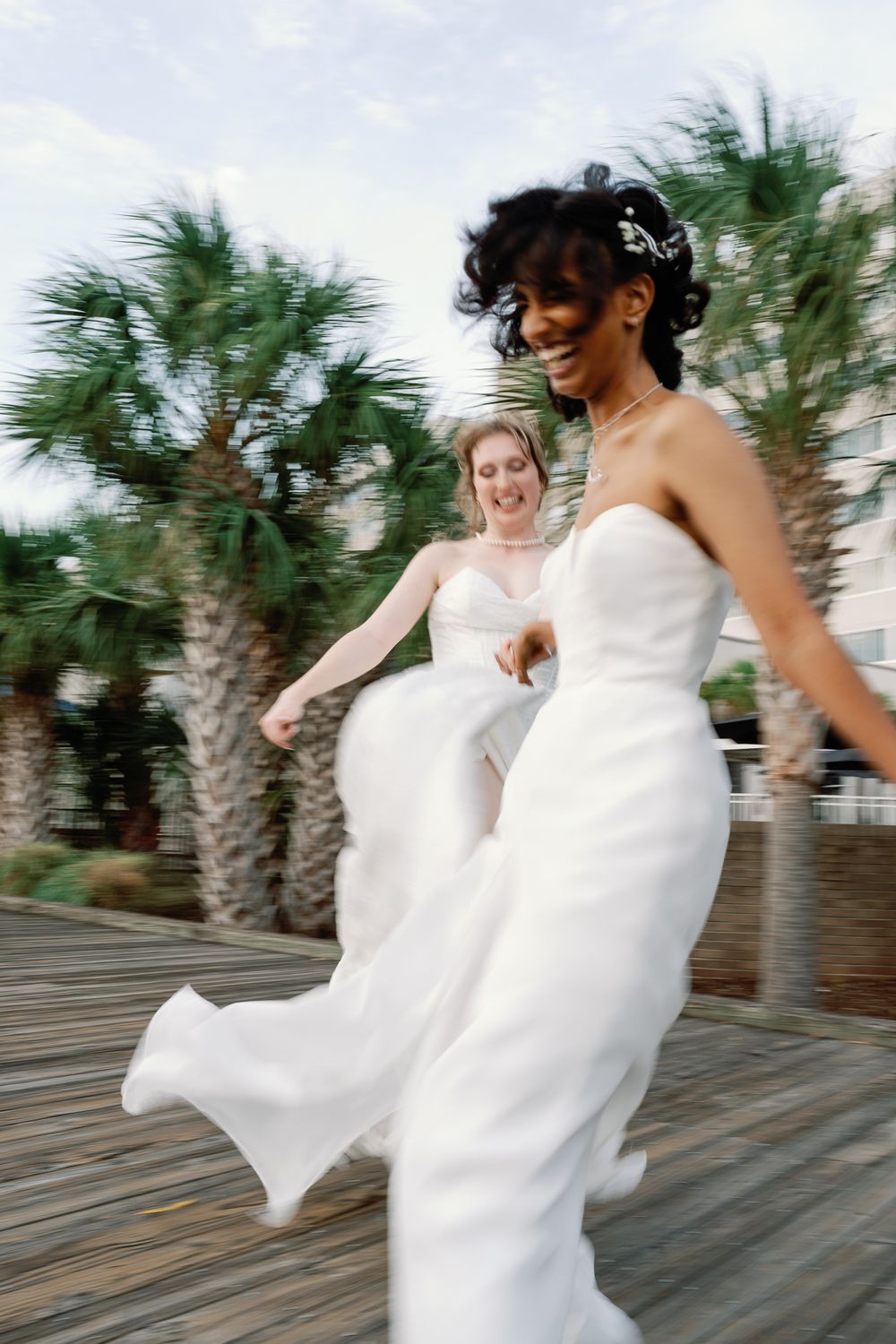 Two brides in white strapless gowns laughing and running on a wooden boardwalk with palm trees.
