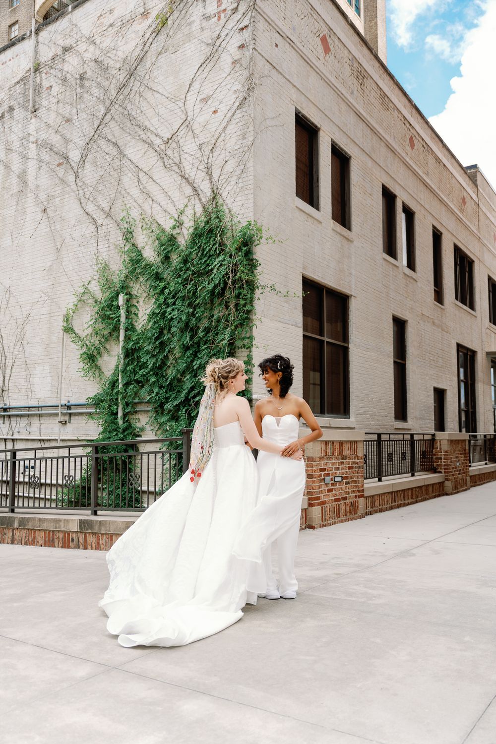 Two brides in white wedding gowns share a romantic moment outside a brick building with ivy-covered walls.