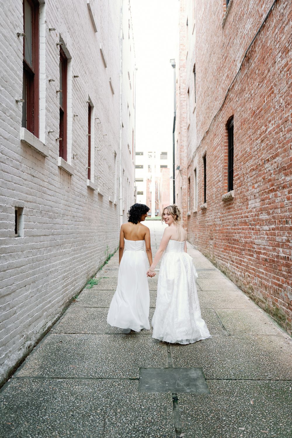 Two brides in white gowns walk hand-in-hand down a narrow brick alley, viewed from behind.