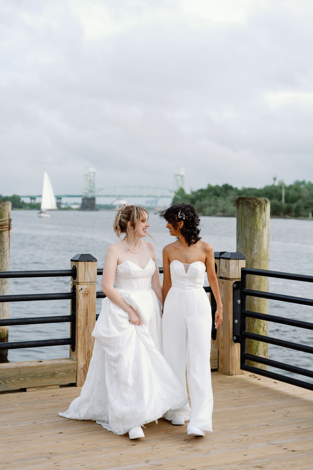 Two brides in white wedding attire walk hand-in-hand on a waterfront dock with a bridge and sailboat in the background.