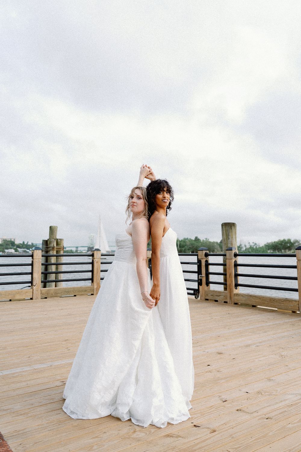 Two brides in white gowns hold hands on a waterfront dock under cloudy skies.