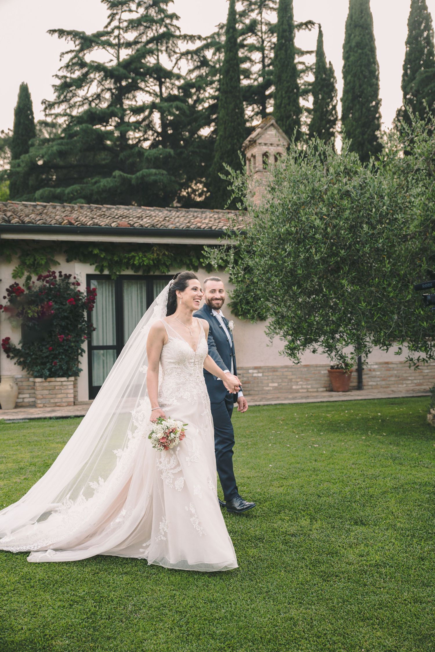 A bride and groom pose in front of an Italian villa with cypress trees.