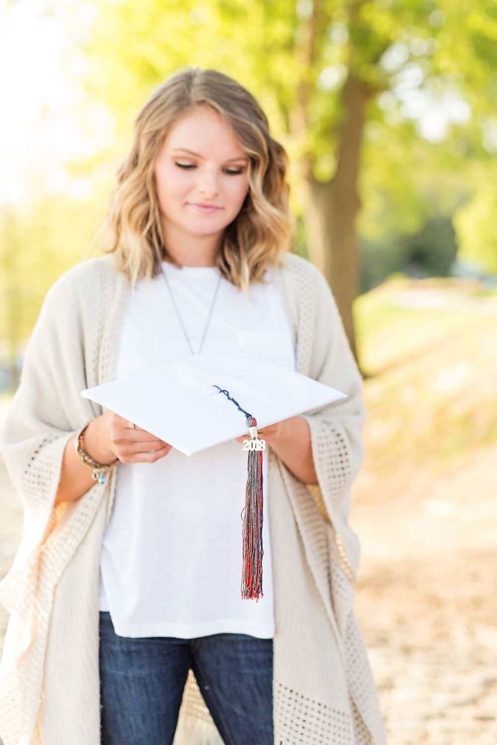 A person holding graduation materials stands outdoors wearing a cream cardigan.