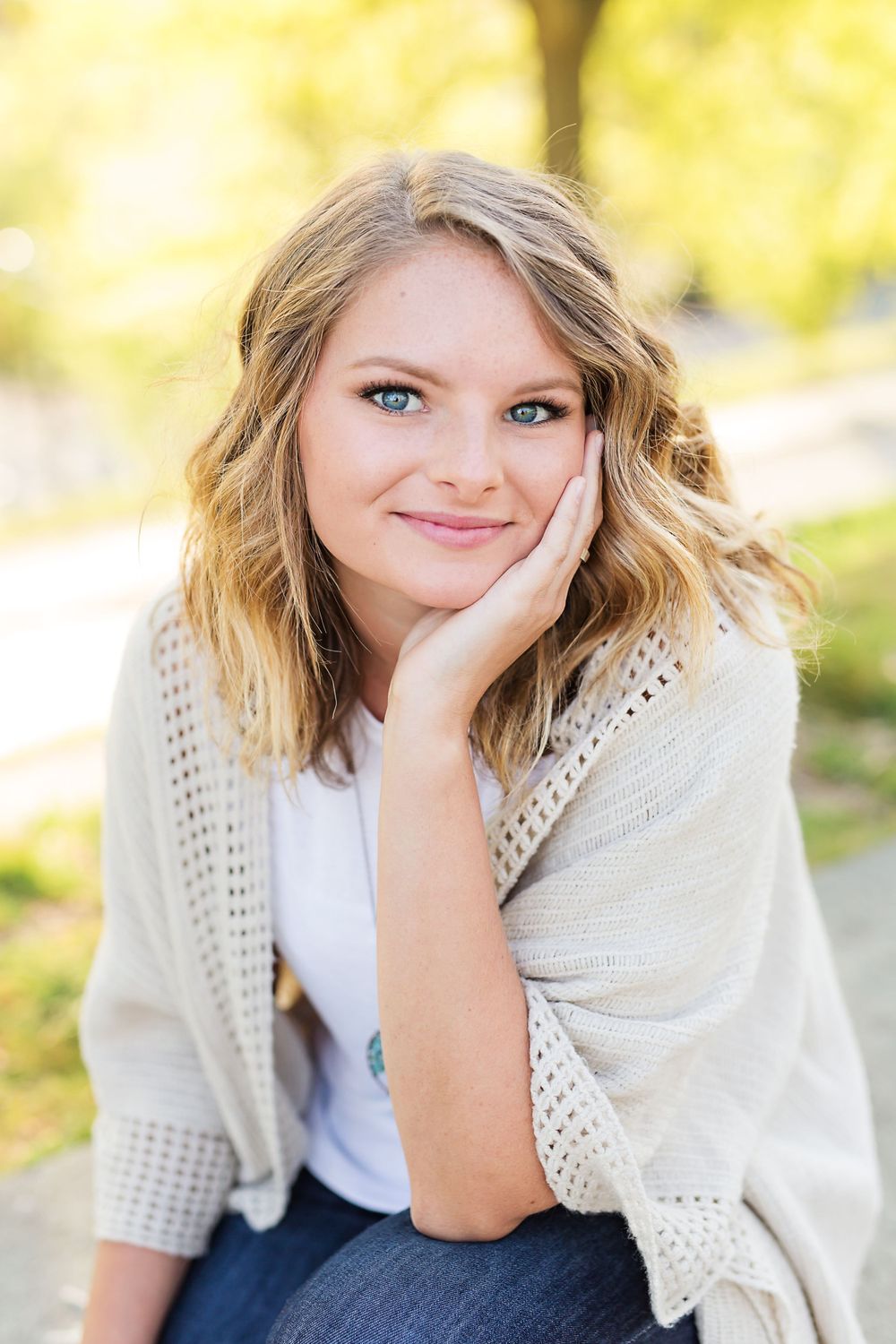A person in a cream sweater poses outdoors with hand resting on cheek.