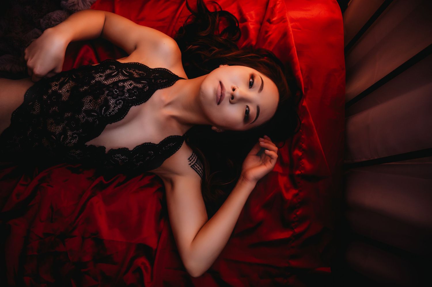 Artistic boudoir photo with black lace lingerie against red satin backdrop in moody lighting.