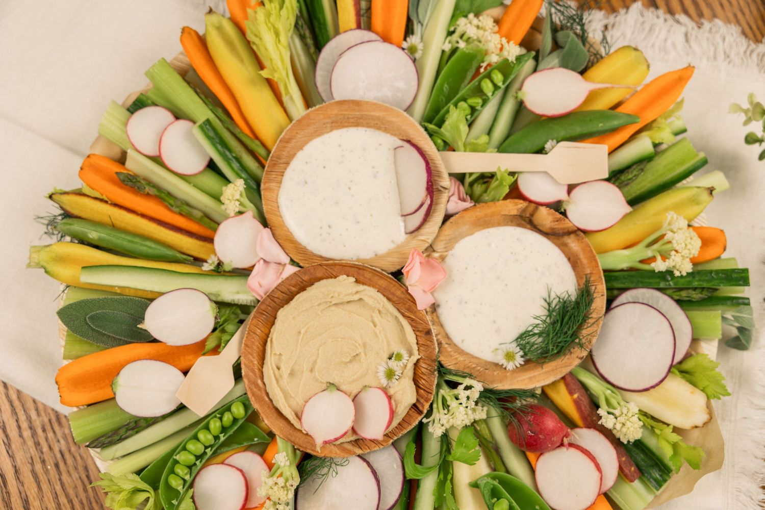 Circular vegetable platter with fresh crudités and various dips in wooden bowls.