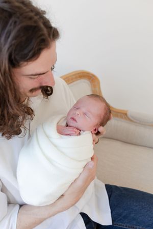 A tender moment as a caregiver cradles a swaddled newborn baby on an elegant cream settee.