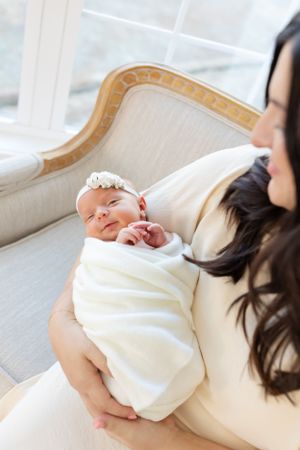 A newborn baby wearing a white headband rests peacefully on a cream-colored settee.