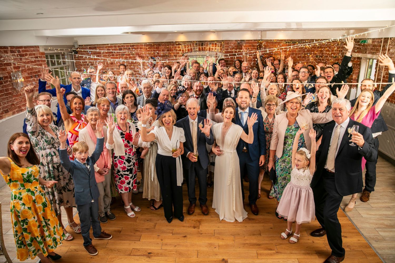 Large group wedding photo with all guests cheering in rustic brick venue.