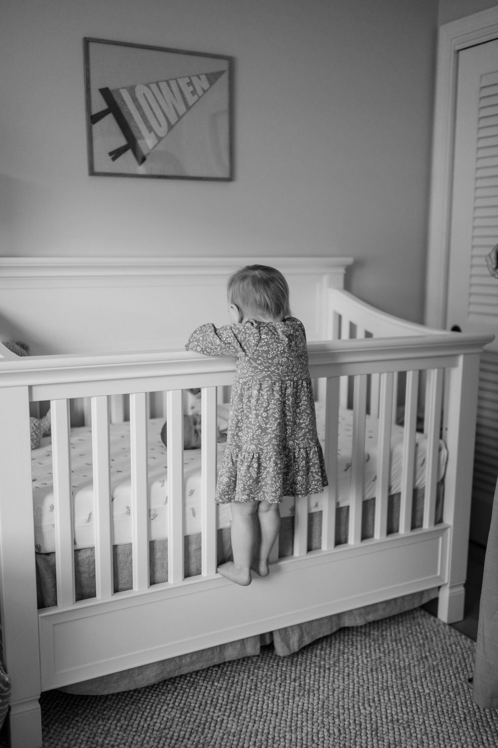 A black and white photo showing a toddler peering over a white crib railing.