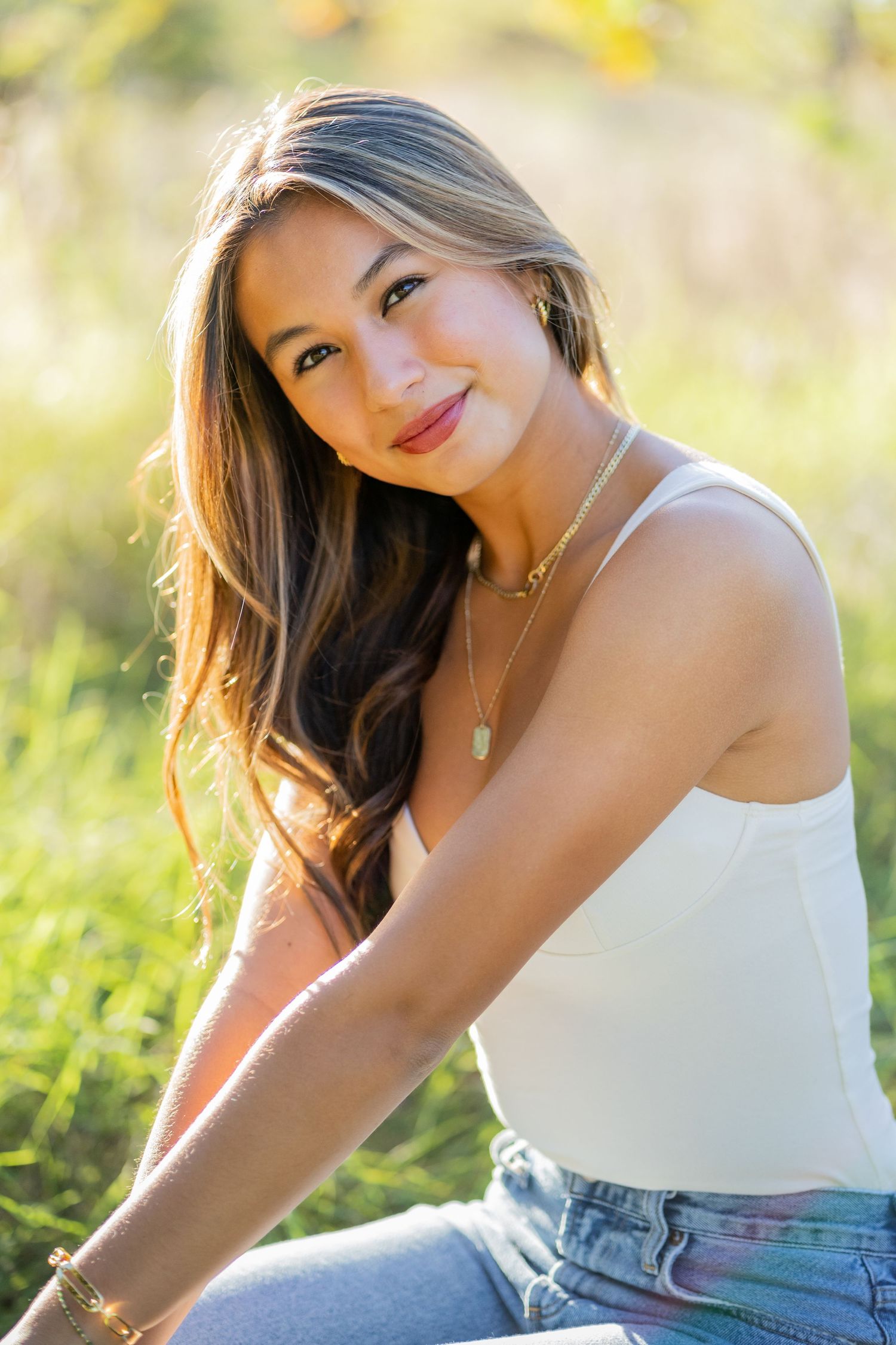 A high school senior in white tank top sitting outdoors in sunlit meadow.