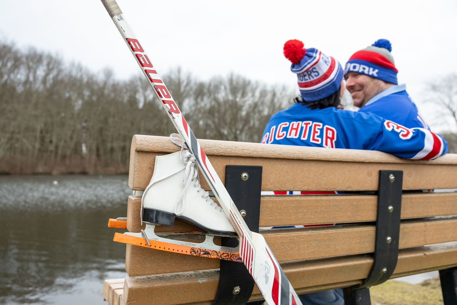 Hockey stick and ice skates rest on wooden bench by frozen pond as couple watches in winter hats.
