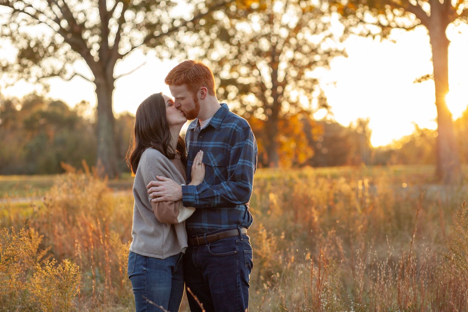 A couple in sweater and flannel shirt kiss in a golden field in Raleigh, NC.
